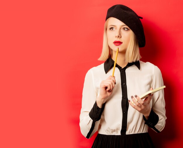 Belle femme française en béret tient un cahier avec un crayon sur un mur rouge