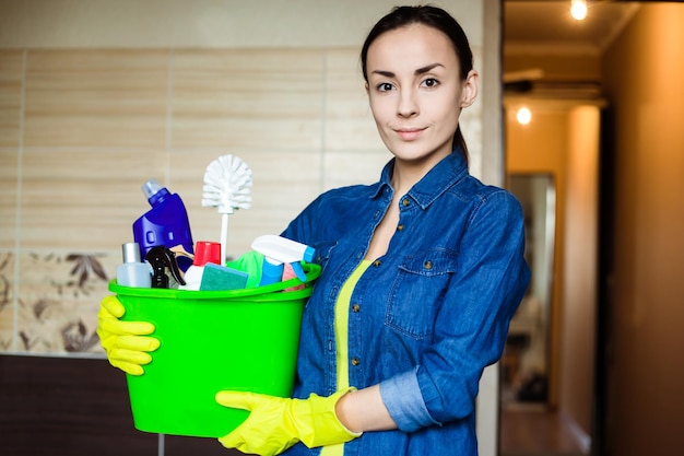 Belle femme en gants de protection tient un seau avec des choses à nettoyer en regardant la caméra et en souriant avant le début du nettoyage de la maison