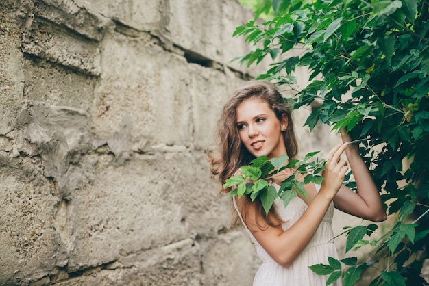 Belle femme heureuse avec des cheveux naturels bouclés en robe blanche près de feuilles d'arbres verts