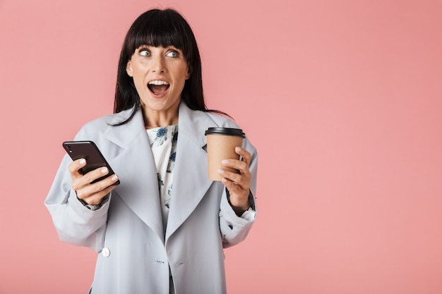 une belle femme heureuse choquée posant isolée sur un mur rose clair à l'aide d'un téléphone portable tenant du café.