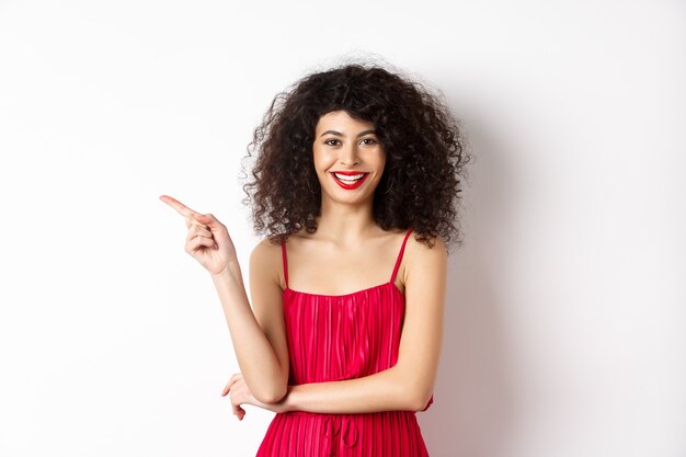 Belle femme heureuse avec une coiffure frisée, vêtue d'une robe de fête, pointant le doigt à gauche sur le logo et souriant, fond blanc.
