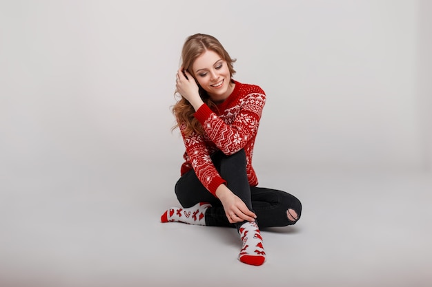 Belle femme heureuse dans un pull en tricot rouge avec une impression de Noël avec des chaussettes assis sur le sol