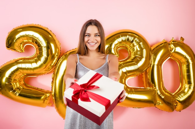 Belle femme heureuse tenant une boîte cadeau rouge devant des ballons de Noël 2020 isolés sur rose