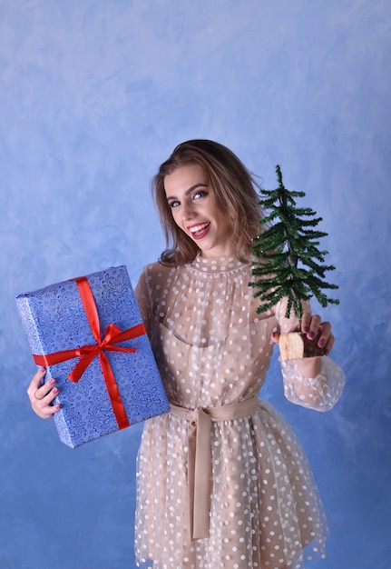 Belle femme heureuse tenant un petit arbre de Noël et un cadeau bleu avec un ruban rouge dans ses mains