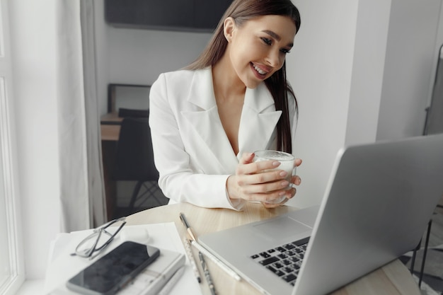 Belle femme heureuse travaillant à la maison avec un ordinateur portable prenant une tasse de café. Femme d'affaires souriante au bureau, à l'aide d'un ordinateur portable