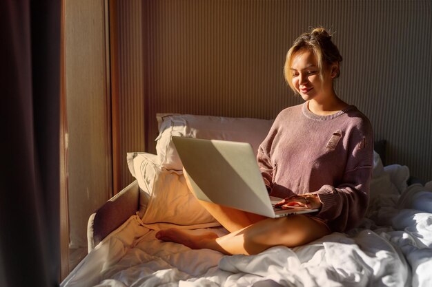 Une belle femme heureuse travaillant sur un ordinateur portable assise sur le lit de la maison.