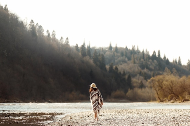 Belle femme hipster marchant sur la plage de la rivière en chapeau de montagne et poncho boho concept de voyage espace pour le texte