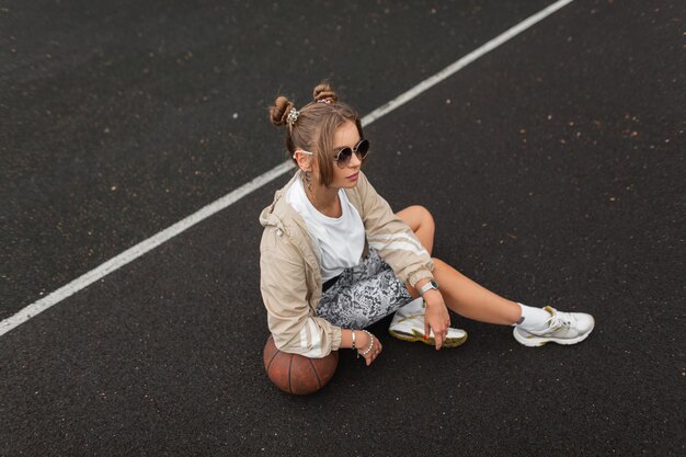 Belle femme hipster sportive à la mode avec des lunettes de soleil dans un short legging coupe-vent à la mode et un t-shirt avec des baskets blanches s'assoit et se repose sur l'asphalte avec une balle