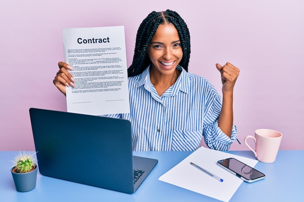 Belle femme hispanique au bureau montrant un document contractuel criant de fierté, célébrant la victoire et le succès très excité avec le bras levé