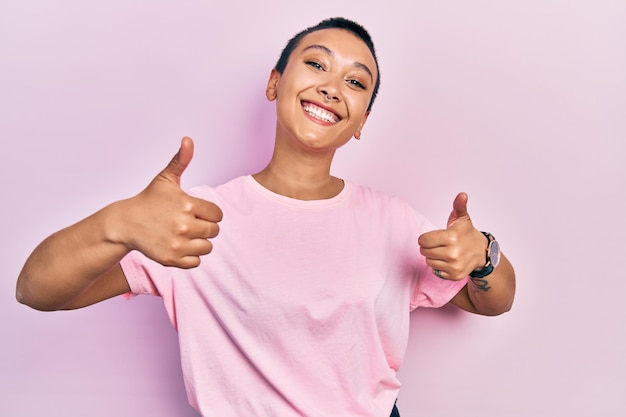 Belle femme hispanique aux cheveux courts portant un t-shirt rose décontracté signe de réussite faisant un geste positif avec la main, les pouces vers le haut souriant et heureux. expression joyeuse et geste gagnant.