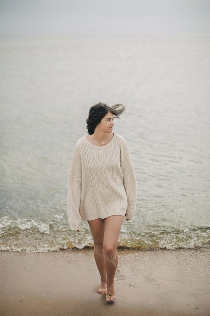 Belle femme insouciante en pull tricoté et avec des cheveux venteux marchant sur une plage de sable à la mer froide