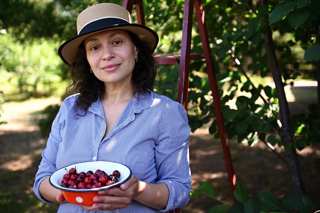 Belle femme jardinier propriétaire d'une ferme écologique montrant des cerises fraîchement cueillies dans son jardin biologique