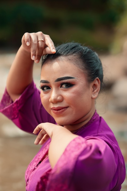 Belle femme javanaise avec maquillage et robe violette posant avec sa main assise sur le rocher près de la rivière