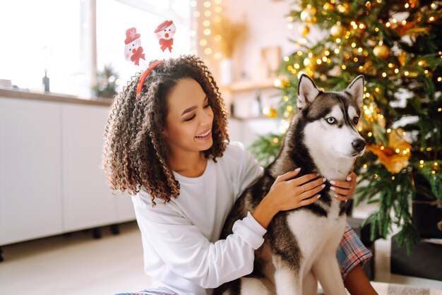 Belle femme jouant et s'amusant avec son chien assis près de l'arbre de noël