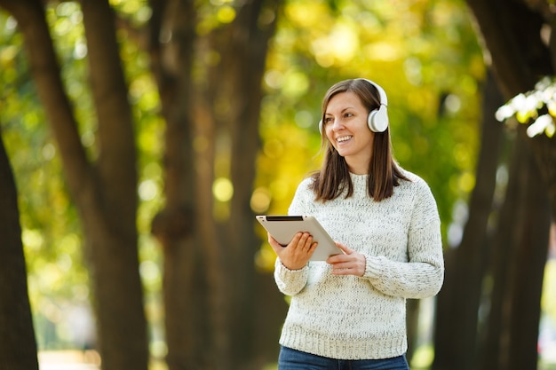 La belle femme joyeuse et joyeuse aux cheveux bruns en pull blanc avec une tablette écoutant de la musique dans les écouteurs blancs dans le parc de l'automne par une chaude journée. Automne dans la ville.