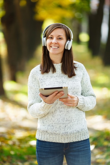 La belle femme joyeuse et joyeuse aux cheveux bruns en pull blanc avec une tablette écoutant de la musique dans les écouteurs blancs dans le parc de l'automne par une chaude journée. Automne dans la ville.