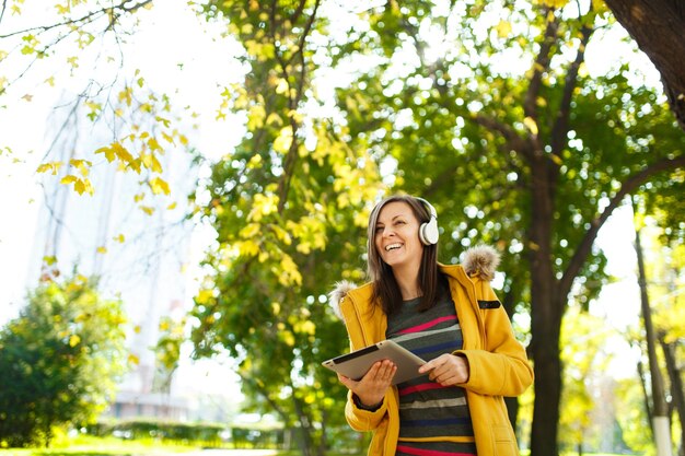 La belle femme joyeuse et joyeuse aux cheveux bruns vêtue d'un manteau jaune et d'une chemise à manches longues rayée se réjouit avec une tablette dans les mains et des écouteurs blancs dans le parc de la ville d'automne par une chaude journée. Feuilles d'or d'automne.