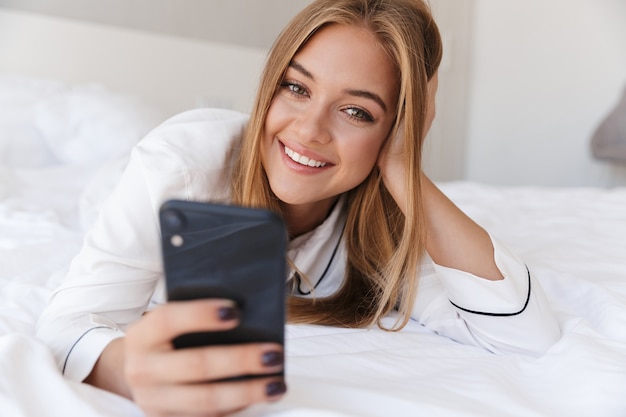 belle femme joyeuse en pyjama utilisant un téléphone portable et souriant en position couchée sur le lit après le sommeil ou la sieste