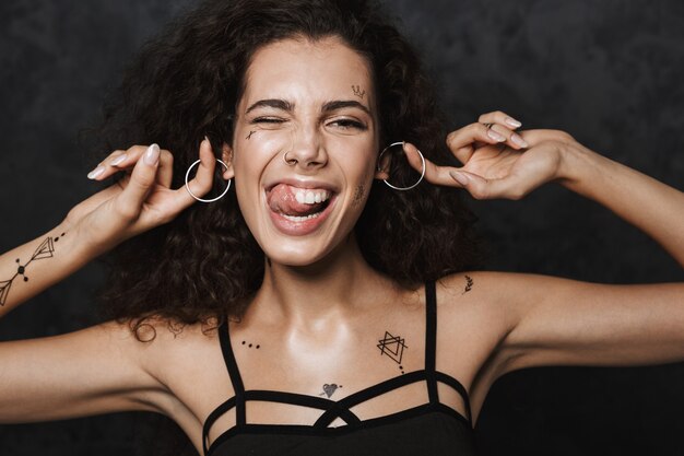 belle femme joyeuse avec un tatouage clignotant et se moquant des boucles d'oreilles isolées sur un mur noir