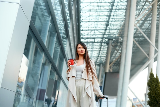 Belle femme joyeuse avec une valise et un café à la main près de l'aéroport