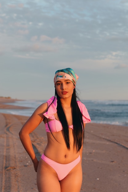 Une belle femme latina portant un bikini rose et un bandana au coucher du soleil posant sur la plage