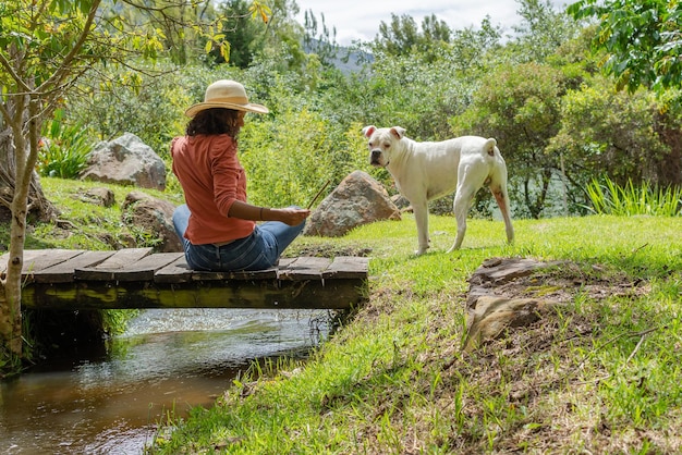 Belle femme latine portant un chapeau assis sur un vieux pont de bois avec un chien