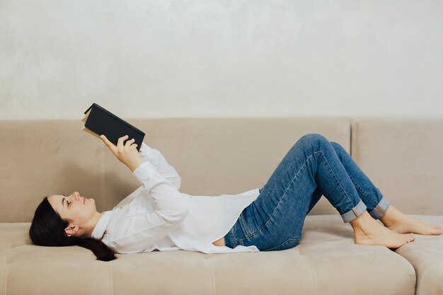 Photo belle femme lisant un livre et allongée sur le canapé dans le salon à la maison.