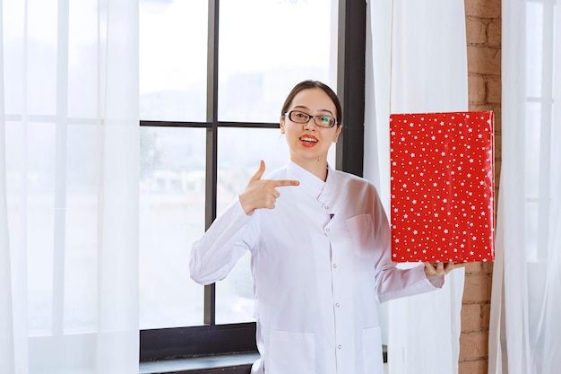 Belle femme avec des lunettes en blouse de laboratoire pointant vers une grande boîte présente près de la fenêtre.