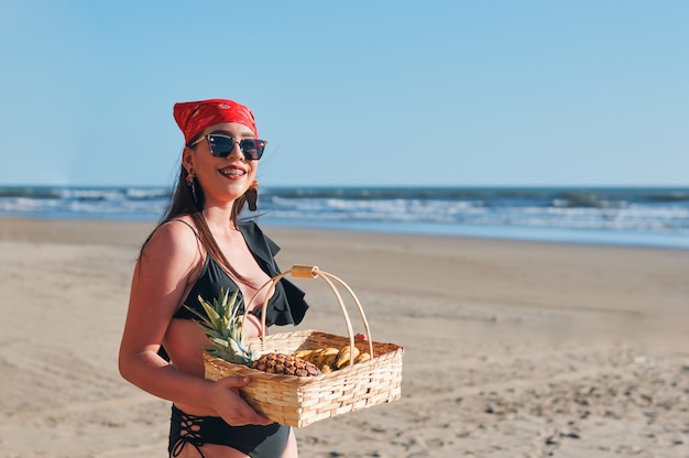 Une belle femme en maillot de bain tenant un panier avec des fruits tropicaux sur la plage