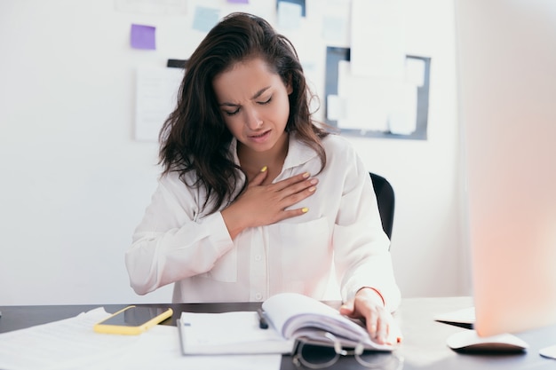 Belle femme malade de ressentir une douleur dans la poitrine tout en travaillant avec des papiers. Main tenant la poitrine après avoir pris un médicament.