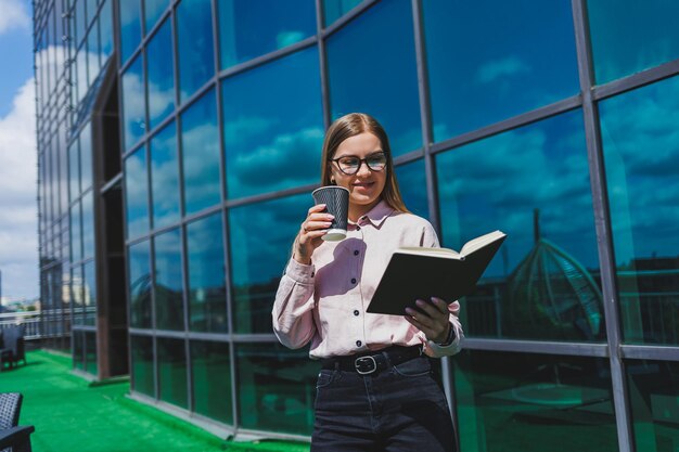 Belle femme manager en lunettes et vêtements à la mode souriant sur la terrasse du bureau la fille boit du café et prend des notes dans un cahier