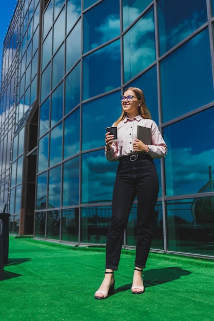 Belle femme manager en lunettes et vêtements à la mode souriant sur la terrasse du bureau la fille boit du café et prend des notes dans un cahier