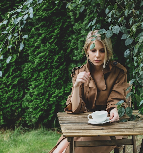 Belle femme en manteau avec une tasse de café à table en bois dans un jardin avec des arbres en arrière-plan