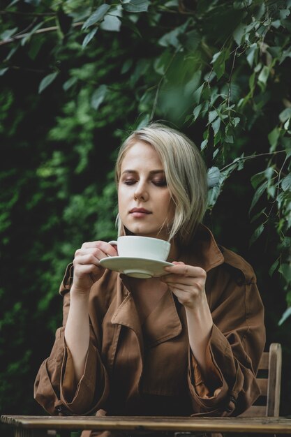 Belle femme en manteau avec une tasse de café à table en bois dans un jardin avec des arbres en arrière-plan