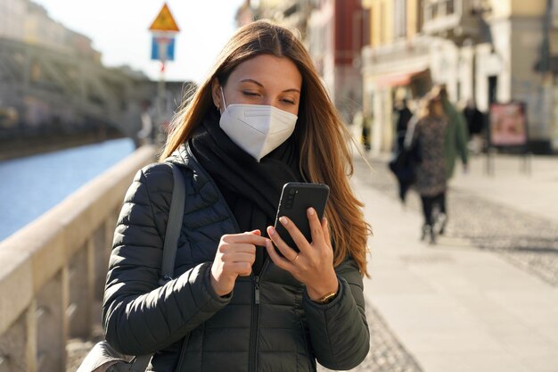 Photo belle femme marchant à l'aide d'un téléphone portable portant un masque facial obligatoire à l'extérieur de la ville en hiver