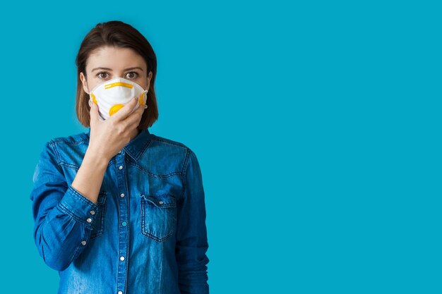 Belle femme avec masque médical et cheveux bruns posant dans une chemise de jeans sur un mur bleu avec un espace vide