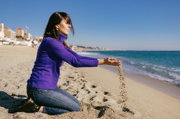 Belle femme mature heureuse jouant avec le sable par une journée d'hiver ensoleillée à la plage