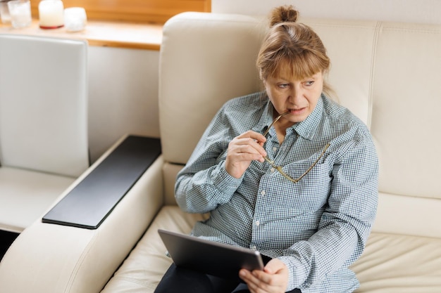 Belle femme mature à lunettes utilise une tablette numérique et souriant assis sur un canapé à la maison