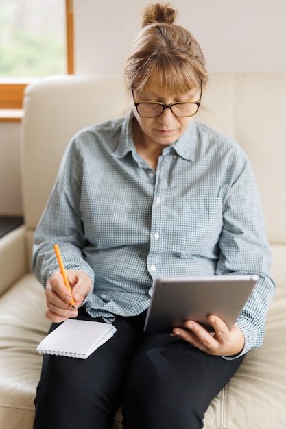 Belle femme mature à lunettes utilise une tablette numérique et souriant assis sur un canapé à la maison