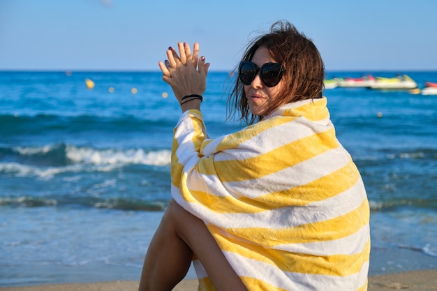 Photo belle femme mature avec une serviette de plage assise sur le sable, une femme souriante heureuse profitant du paysage de coucher de soleil sur la mer. détente, loisirs, beauté, océan, mode de vie des personnes d'âge moyen