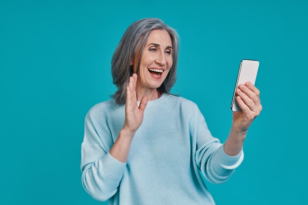 Belle femme mature utilisant un téléphone intelligent et souriant en se tenant debout sur fond bleu