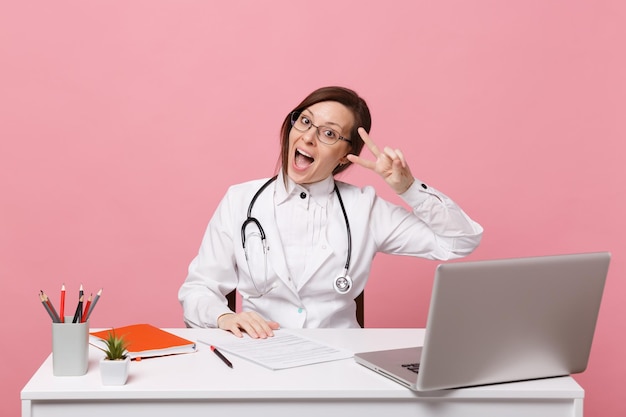 Belle femme médecin assise au bureau travaille sur ordinateur avec un document médical à l'hôpital isolé sur fond de mur rose pastel. Femme en robe médicale lunettes stéthoscope. Concept de médecine de santé