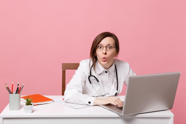 Belle femme médecin assise au bureau travaille sur ordinateur avec un document médical à l'hôpital isolé sur fond de mur rose pastel. Femme en robe médicale lunettes stéthoscope. Concept de médecine de santé