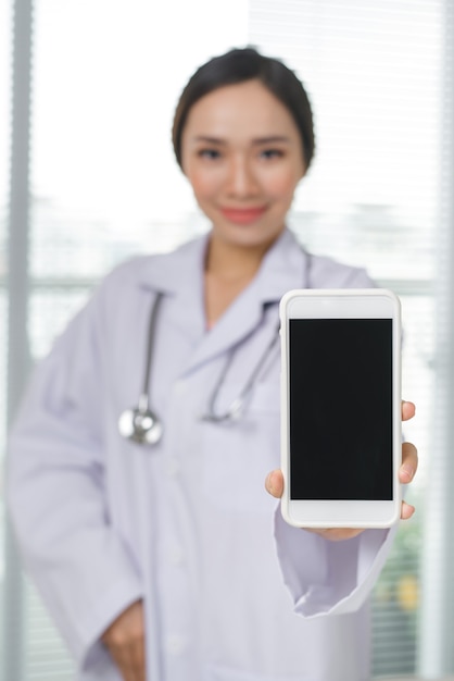 Une belle femme médecin souriante avec un stéthoscope montrant l'écran du smartphone sur fond intérieur de la clinique.