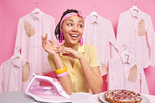 Belle femme de ménage rêveuse à la peau foncée regarde de côté, sourit joyeusement, fait des travaux ménagers et des poses de lessive près d'une planche à repasser avec une tarte au fer électrique qui travaille dur à la maison