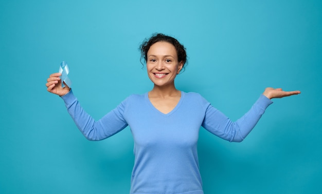 Une Belle Femme Métisse Souriante Portant Des Vêtements Décontractés Bleus, Tient Un Ruban De Sensibilisation Bleu Et Met La Paume En L'air Tenant Un Espace De Copie Imaginaire Pour La Publicité Médicale Pour La Journée Mondiale Du Diabète, Le 14 Novembre