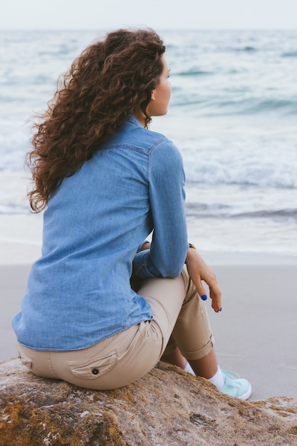 Belle femme mince aux cheveux bouclés, assise sur un rocher au bord de la mer