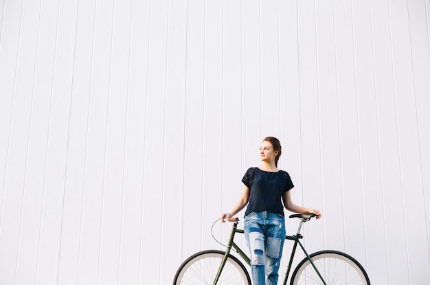 Belle femme à la mode debout avec vélo, en détournant les yeux, sur le mur blanc. Copiez l'espace. En plein air