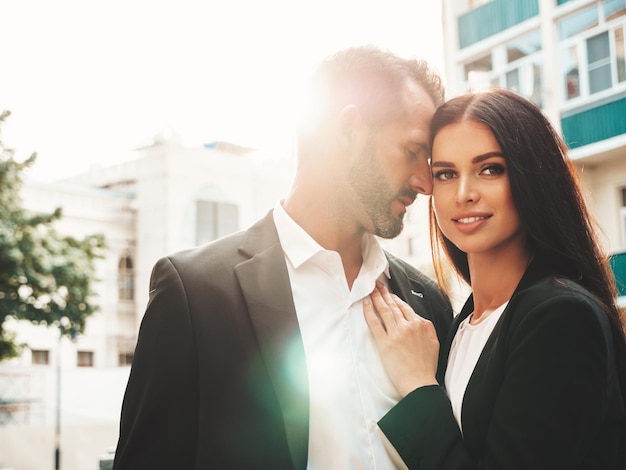 Belle femme de mode et son beau petit ami en costume Sexy modèle brune souriante en robe de soirée noire Couple à la mode posant dans la rue au coucher du soleil Homme brutal et sa femme à l'extérieur