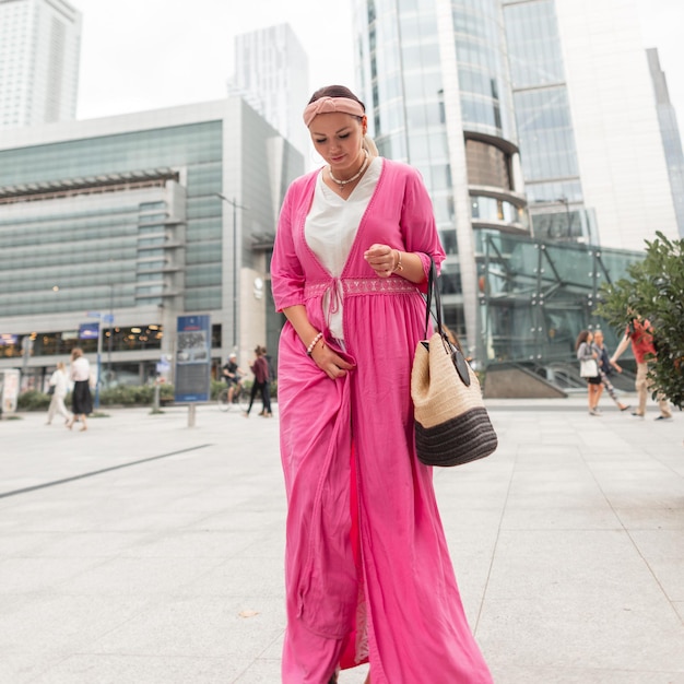 Belle femme à la mode en vêtements d'été dans un paréo rose élégant avec un sac en osier de mode se promène dans la ville